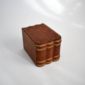 May include: A brown leather playing card box with a book-like design. The box features gold trim and decorative card suit symbols on the top. The edges are rounded, and the box appears to be closed.