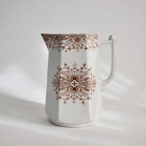 May include: White ceramic pitcher with a brown floral pattern. The pitcher has a handle and a spout. The floral design is symmetrical and covers the top and center of the pitcher. The pitcher has a geometric shape.