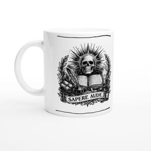 Op de afbeelding: Witte keramische mok met een zwart-wit illustratie. Het ontwerp bevat een schedel, een open boek, een veer en de tekst "SAPERE AUDE" op een banner. De mok heeft een gebogen handvat.