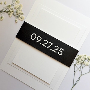 May include: A stack of white paper cards with a black band across the center. The band displays the date "09.27.25" in white. Delicate white flowers are arranged near the cards, creating a minimalist and elegant design.