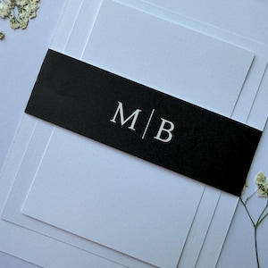 May include: A stack of white cards with a black band featuring the initials "M|B" in a serif font. The cards are layered, creating a sense of depth. Delicate white flowers are scattered around the edges, adding a touch of elegance.
