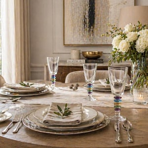 Op de afbeelding: Een prachtig gedekte eettafel met witte borden, bestek en elegant glaswerk. De glazen hebben kleurrijke kralen stelen. Een vaas met witte bloemen en brandende kaarsen dragen bij aan de sfeer. De tafel is gedekt voor een speciale gelegenheid.