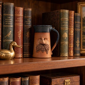 May include: A ceramic mug with a dark blue glazed rim and a natural terracotta body featuring a sculpted face with a prominent nose and a textured mustache. The mug is positioned on a shelf between several antique hardcover books.