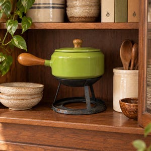 May include: A bright lime green fondue pot with a wooden handle and matching lid knob, resting on a black metal stand. The pot features a smooth, glossy finish and is positioned on a wooden shelf alongside ceramic bowls and wooden spoons.