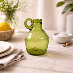 May include: A small, green glass pitcher with a handle. The pitcher has a crackled texture and a rounded body. The background includes a basket with a lemon, plates, and a small bowl with white contents.