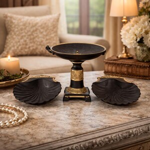 May include: A decorative centerpiece with a black bowl on a pedestal, flanked by two black shell-shaped dishes. The pedestal has gold accents. A lit candle and a pearl necklace are also visible.