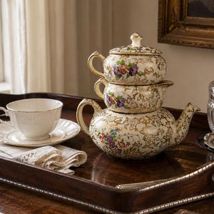May include: A stackable tea set featuring a teapot, creamer, and lidded sugar bowl in cream ceramic with gold floral filigree and colorful flower accents. Beside it sits a plain white teacup and saucer, a lace napkin, and a clear glass sugar jar.