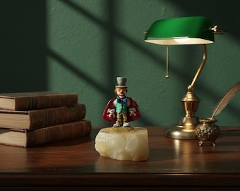 May include: A decorative figurine of a circus ringmaster wearing a red cape with white dog figures, a top hat, and a blue bow tie. The figurine is displayed on a light-colored stone base. A green desk lamp and antique books are in the background.