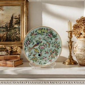May include: A decorative plate with a light green background and a floral and bird design. The plate is displayed on a clear stand. A gold candlestick with a lit candle and a vase with dried flowers are also visible.