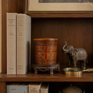 May include: A cylindrical container with a brown and orange finish, featuring a repeating geometric Greek key pattern and floral motifs, resting on a dark carved wooden stand. The container is positioned between two beige hardcover books.