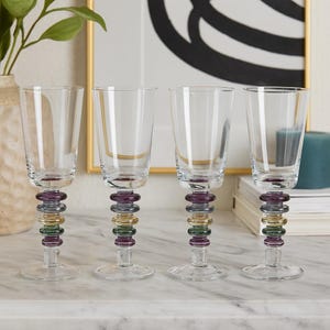 Peut inclure: Quatre verres à vin transparents avec des tiges décoratives. Les tiges présentent un design empilé d'anneaux violets, dorés et verts. Les verres sont exposés sur une surface blanche, avec un arrière-plan flou.