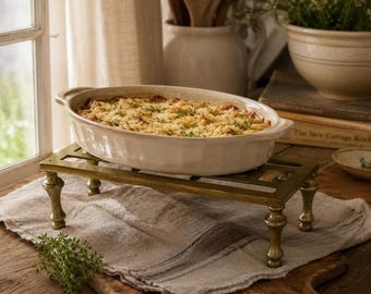 Può includere: Una pirofila ovale in ceramica color crema riempita con un pasticcio dorato al forno guarnito con erbe verdi. Il piatto poggia su un sottopentola rettangolare in metallo color ottone con quattro gambe decorative e una superficie a griglia.