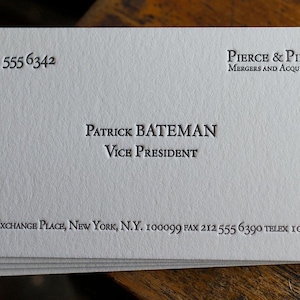 May include: A stack of white business cards with embossed black text. The card displays the name "Patrick Bateman", title "Vice President", and contact information for "Pierce & Pierce Mergers and Acquisitions" in New York City.