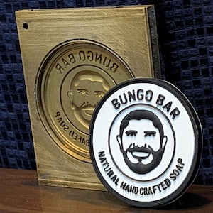 Puede incluir: Primer plano de un sello metálico dorado y un jabón redondo en blanco y negro. El sello tiene las palabras "BUNGO BAR" y un diseño de rostro. El jabón dice "BUNGO BAR NATURAL HAND CRAFTED SOAP".