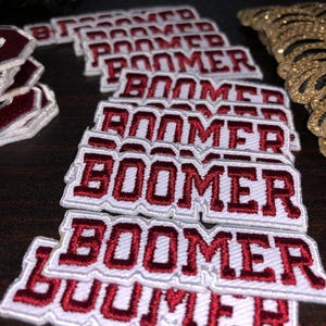 Puede incluir: Un conjunto de parches bordados con la palabra "BOOMER" en letras granates sobre un fondo blanco. Los parches están apilados y dispuestos sobre una superficie oscura, con parches adicionales y adornos de lentejuelas doradas visibles.