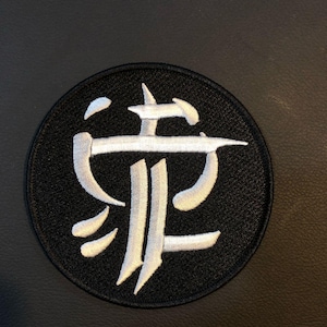 May include: Circular black embroidered patch with a white, stylized symbol. The symbol has a central vertical line with horizontal and curved lines. The patch is designed for clothing or accessories.