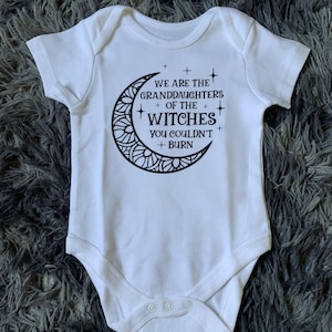 Puede incluir: Body de bebé blanco con mangas cortas. El body presenta un diseño de media luna negra con el texto "WE ARE THE GRANDDAUGHTERS OF THE WITCHES YOU COULDN'T BURN". Tiene tres cierres de presión en la parte inferior.