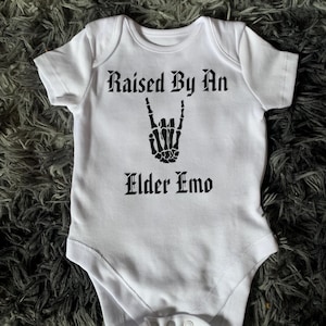 May include: A white short-sleeved baby bodysuit featuring black text that reads Raised By An Elder Emo. Between the two lines of text is a black graphic of a skeleton hand making a rock and roll hand gesture with the index and pinky fingers extended.