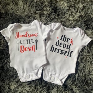 Puede incluir: Dos bodies blancos para bebé. Uno tiene el texto "Handsome LITTLE Devil" en rojo y negro. El otro tiene el texto "the devil herself" en negro y rojo. Ambos bodies tienen mangas cortas y cierres de broche.
