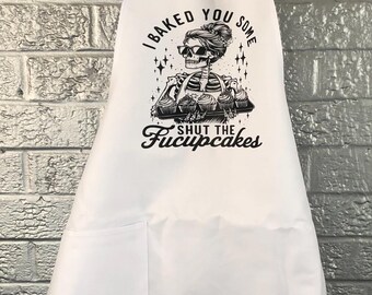 Op de afbeelding: Witte schort met een grote voorzak en een nekband. De schort heeft een zwart-witte afbeelding van een skelet met cupcakes, met de tekst "I baked you some shut the Fucupcakes."