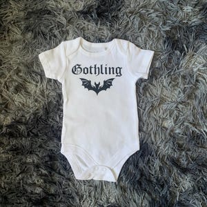 以下が含まれることがあります： 黒いゴシックフォントで「Gothling」の文字と黒いコウモリのグラフィックがプリントされた白いベビーボディスーツ。半袖で、下部にスナップ留めが付いています。グレーと白のテクスチャード加工された表面に置かれています。
