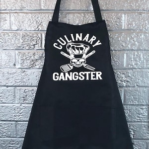 May include: Black apron with the text "CULINARY GANGSTER" in white. The design includes a chef's hat, crossed utensils, and a skull. The apron has a neck strap and a large front pocket.