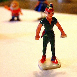 May include: A green and red plastic figurine of Peter Pan standing on a white base. He is wearing a green shirt and pants with a red hat and red shoes.