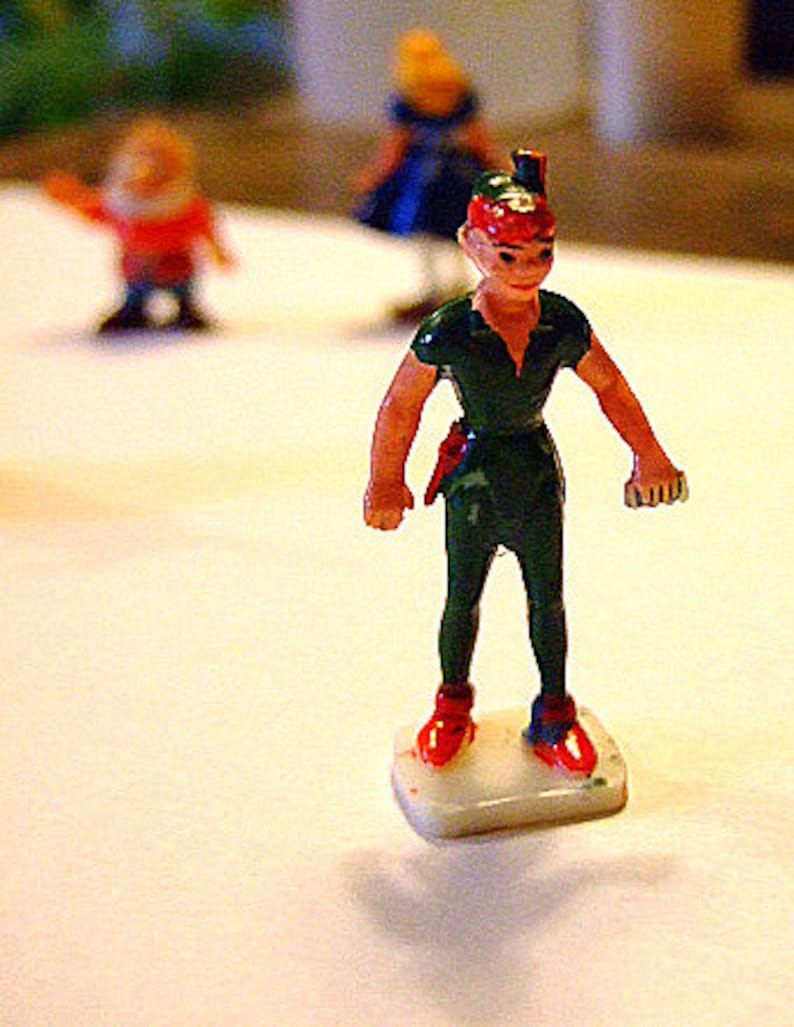 May include: A green and red plastic figurine of Peter Pan standing on a white base. He is wearing a green shirt and pants with a red hat and red shoes.