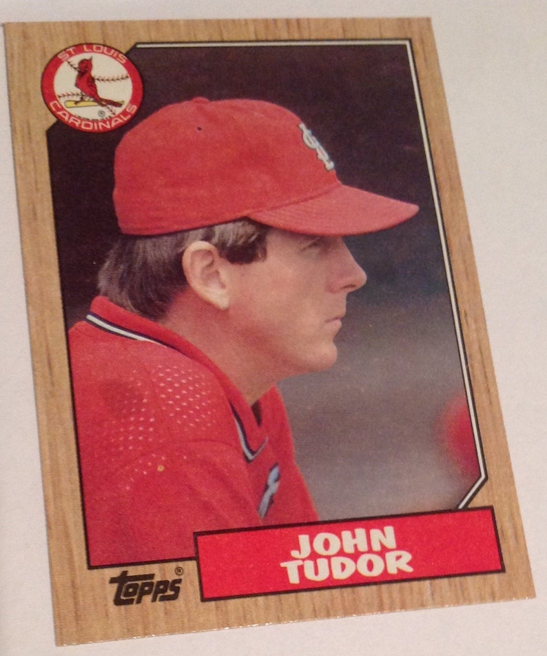 Puede incluir: Una tarjeta de b&eacute;isbol con un retrato de John Tudor, un ex lanzador de la Major League Baseball. La tarjeta es roja, blanca y azul con el texto "John Tudor" y el logotipo de Topps. La tarjeta presenta una foto de Tudor con una gorra y una camiseta rojas de los St. Louis Cardinals.