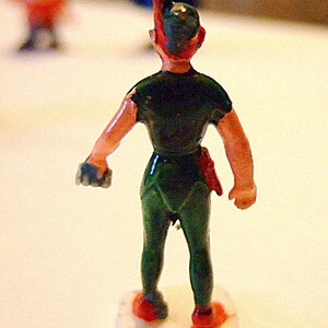 May include: A green and red plastic toy figurine of Peter Pan, standing on a white base. He is wearing a green tunic and pants, and has a red hat with a feather.