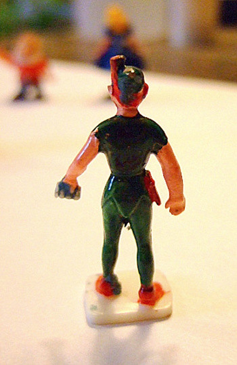 May include: A green and red plastic toy figurine of Peter Pan, standing on a white base. He is wearing a green tunic and pants, and has a red hat with a feather.