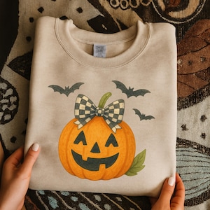 Può includere: Felpa beige con una zucca arancione sorridente, un fiocco a scacchi e tre pipistrelli. La felpa girocollo ha un'etichetta "Comfort Colors". Un divertente design a tema Halloween.