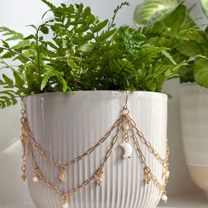 May include: A white ceramic plant pot with a fern, decorated with a gold chain and pearl details. The chain includes a crescent moon charm and hanging beads. The pot has a ribbed texture.