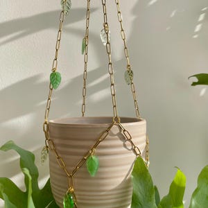 May include: A beige ceramic planter with horizontal ridges suspended by a gold-toned metal chain hanger. The chain features small, translucent green and clear glass leaf-shaped charms attached at various intervals along the links.