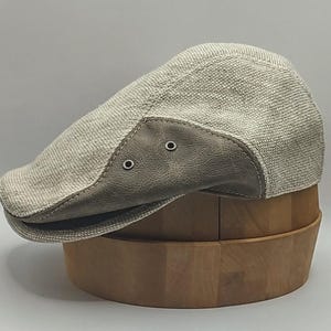 Könnte beinhalten: Eine hellbeige Newsboy-Mütze mit einem grau lederartigen Akzent am Schirm. Die Mütze hat zwei kleine Ösen an der Seite des Schirms. Die Mütze ist aus einem gewebten Stoff gefertigt und wird auf einem Holzständer präsentiert.
