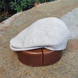 Puede incluir: Gorra plana gris claro de tela texturizada, sobre una horma de sombrero de madera marrón. La gorra tiene una corona redondeada y una pequeña visera curva. El fondo presenta una superficie de piedra y hojas verdes.