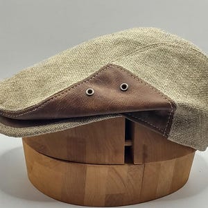Könnte beinhalten: Eine beige Newsboy-Cap mit braunen Lederdetails an Krempe und Seiten. Die Cap besteht aus einem strukturierten Stoff und hat zwei kleine Metallösen am Leder. Sie wird auf einem hölzernen Hutständer präsentiert.