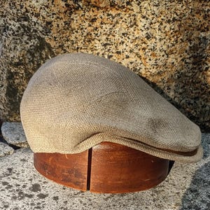 Könnte beinhalten: Eine beigefarbene Newsboy-Cap mit braunem Lederbesatz, die auf einem hölzernen Hutständer liegt. Die Cap hat ein strukturiertes Gewebe und ein klassisches, abgerundetes Design. Im Hintergrund ein großer, strukturierter Felsen.
