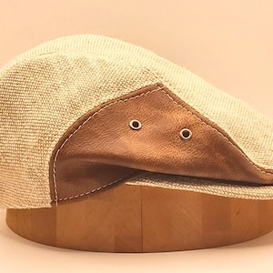 Könnte beinhalten: Beige Newsboy-Cap mit braunem Leder-Visier. Die Kappe besteht aus einem gewebten Stoff und hat zwei kleine Metallösen am Visier. Der Hut wird auf einer Holzform präsentiert.
