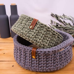 Puede incluir: Dos cestas de ganchillo hechas a mano, una gris y otra verde oliva, están apiladas sobre una superficie de madera. La cesta gris es más grande, con una etiqueta de cuero y un botón dorado. La cesta verde oliva tiene una etiqueta similar. Dos botellas gris oscuro en el fondo.