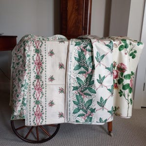 May include: Assortment of floral patterned fabric rolls displayed on a wooden cart. The fabrics feature designs with pink ribbons, green leaves, and red roses on a cream background. The cart has a dark wooden wheel.