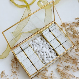 May include: A rectangular glass box with a gold-colored metal frame, filled with white candies and small chocolate bars. The chocolate bars are labeled "EMMA & NOAH". A gold ribbon and dried flowers are arranged around the box.