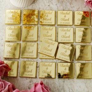 Puede incluir: Un conjunto de pequeños cuadrados dorados, grabados con los nombres "Rachel & Mark" y la fecha "31.10.2025". Estos recuerdos, dispuestos sobre una superficie clara, son ideales para bodas o eventos especiales.