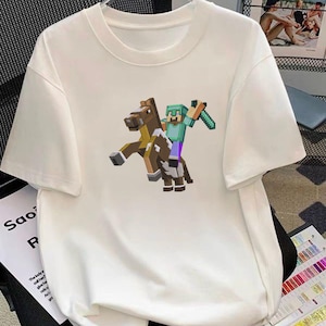 May include: White t-shirt featuring a pixelated graphic of a character riding a horse, holding a sword. The character wears a green helmet and blue shirt. The horse is brown and white. The shirt has a crew neck and short sleeves.