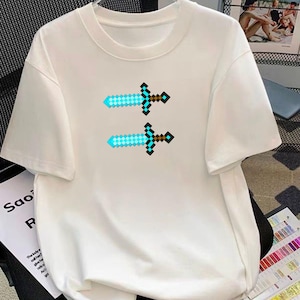 May include: White t-shirt featuring two pixelated swords with blue and brown details. The swords are arranged vertically, creating a symmetrical design. The shirt has a crew neck and short sleeves.