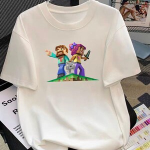 Könnte beinhalten: Weißes T-Shirt mit einem farbenfrohen Grafikdesign von zwei blockartigen Figuren und einem Wolf auf einer pixeligen grünen und braunen Landschaft. Eine Figur zeigt, die andere hält ein Schwert. Das Shirt hat kurze Ärmel und einen Rundhalsausschnitt.