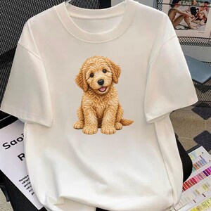 May include: Cream-coloured t-shirt with a detailed illustration of a golden-brown puppy. The puppy is sitting, with its tongue slightly out. The shirt has short sleeves and a crew neck. The text "Saoi R" is partially visible on the left side.