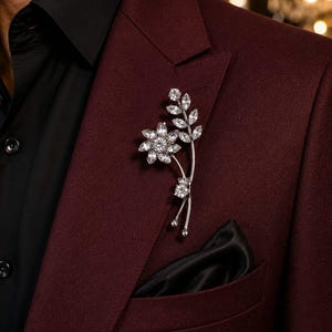 以下が含まれることがあります： 花と葉の模様に配置された透明なファセットカットクリスタルをあしらったシルバー調のフラワーブローチ。ダークバーガンディのブレザーに留められており、その下には黒いシャツを着用し、胸ポケットには黒いポケットチーフが挿されています。