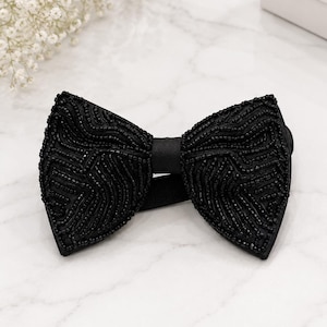 May include: A black beaded bow tie with a satin centre. The bow tie is heavily embellished with small, dark beads, creating a textured surface. The background is a marble-patterned surface.