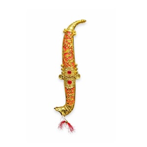 May include: Ornate gold dagger with a red handle. The handle features intricate floral patterns and is adorned with three red gemstones. A red tassel hangs from the base of the dagger, adding a decorative touch. The dagger is set against a white background.